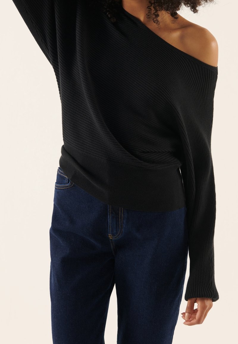 Femme portant un pull côtelé noir à épaules dénudées et un jean bleu foncé, un bras levé et des cheveux bouclés visibles.