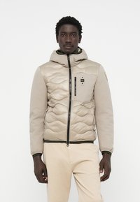 Beige gevoerde jas met capuchon, voorzien van een zwarte rits, groene accenten en een borstzak. Gladde stof met een gestructureerd patroon.