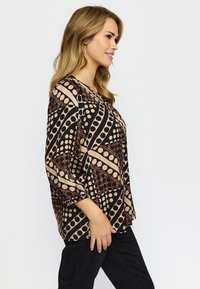 Kvinna med blont hår som bär en beige och svart blus med prickigt mönster och svarta byxor, stående i profil mot en enfärgad bakgrund.