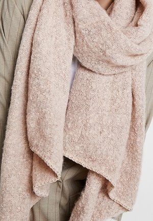 Écharpe douce et texturée couleur rose clair, enveloppée autour du cou et des épaules, portée sur une veste de costume beige à fines rayures.