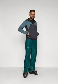 Férfi kültéri öltözék sötét türkizkék softshell kabáttal világoskék részletekkel és sötét türkizkék nadrággal; a kabát cipzárral záródik, a nadrágnak cipzáras zsebei vannak.