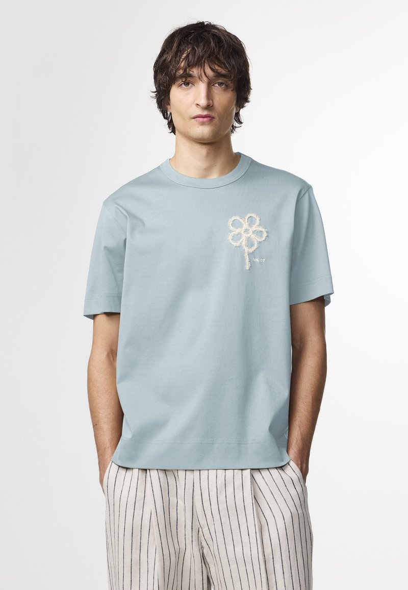 Lyseblå bomulds t-shirt med et tekstureret blomsterdesign i creme på brystet og korte ærmer, parret med stribede beige bukser.