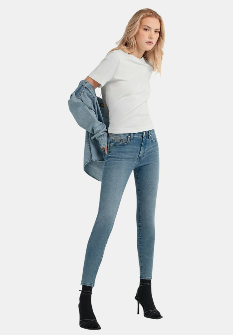Top corto bianco aderente a maniche corte, jeans skinny blu chiaro e un giubbotto di jeans appoggiato sulle spalle. Scarpe nere con tacco.