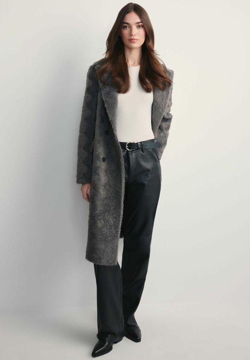 Szary płaszcz z sztucznego futra o podwójnym zapięciu, zestawiony z czarnymi skórzanymi spodniami i dopasowaną białą bluzką. Elegancki czarny pasek w akcentach.