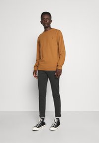 Brun langærmet skjorte, grå tætsiddende bukser og sorte højtop sneakers. Skjorten har et lille logo på brystet. Enkel og strømlinet design.