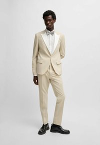 Abito beige su misura con colletto a scialle bianco, pantaloni coordinati e papillon grigio chiaro. Scarpe nere completano il look. Finitura in tessuto liscio.