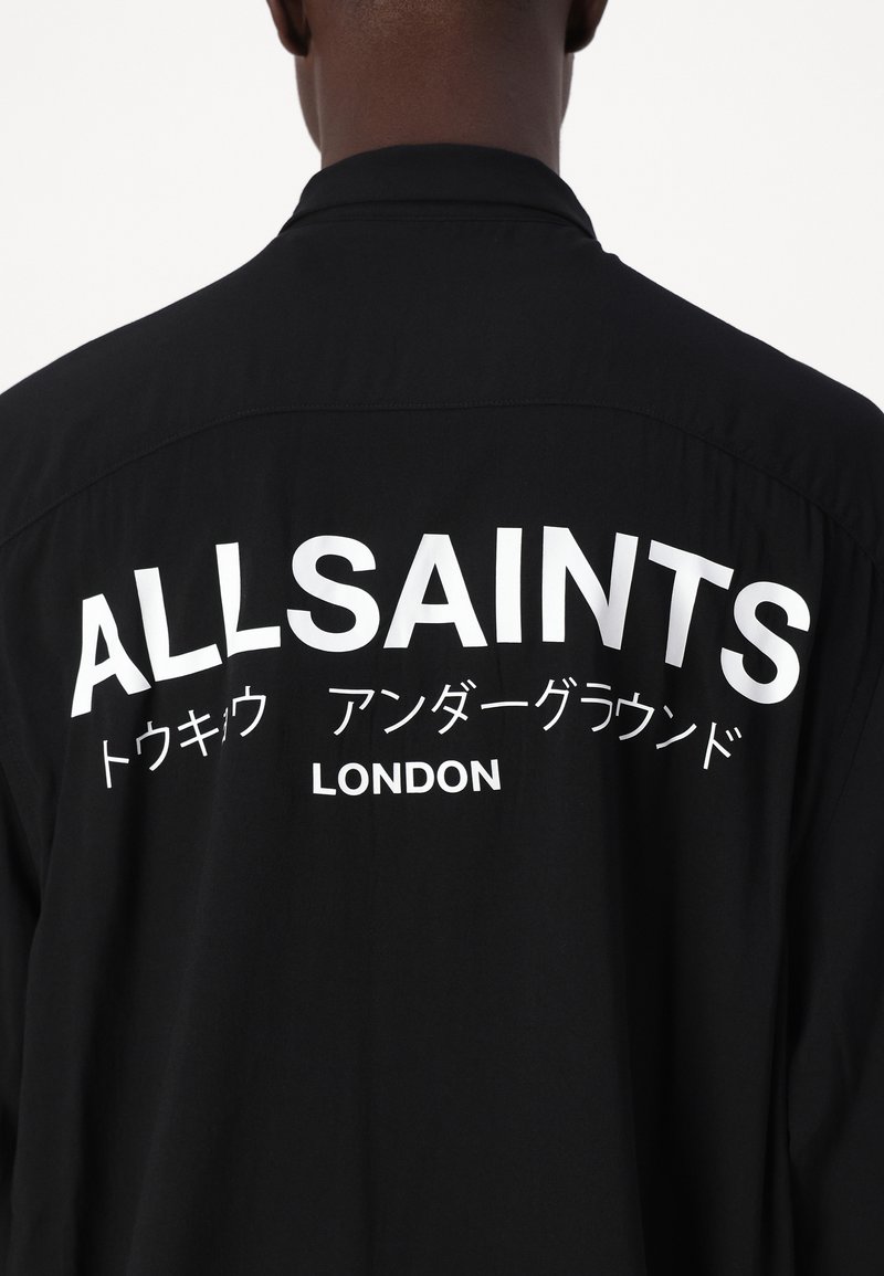 Zwarte shirt met grote witte tekst op de achterkant die "ALLSAINTS" leest, met daaronder aanvullende kleine tekst in het Japans en "LONDON".