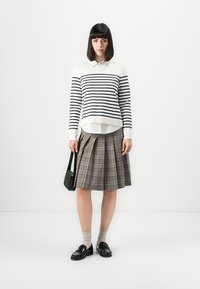 Gestreifter Creme- und Marinepullover über einem weißen Hemd; gefalteter grau-karierter Rock; schwarze Loafers; Halten einer schwarzen Handtasche; beigefarbene Socken.