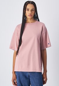 Camiseta de algodón rosa con un corte holgado, escote redondo y mangas cortas. Destaca el logo en la manga. Combinada con jeans de denim azul.