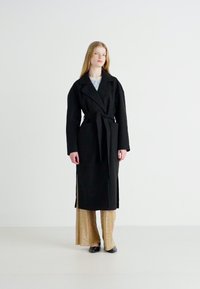 Μαύρο μακρύ παλτό με γιακά με πέτα, ζώνη στη μέση, πλαϊνές σχισμές και υφή, σε συνδυασμό με πλισέ χρυσά παντελόνια.