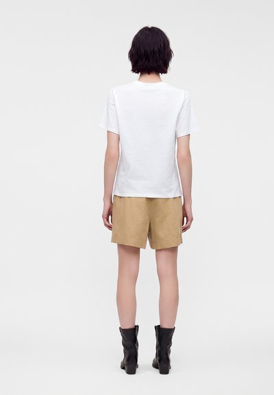 Personne aux cheveux courts et foncés porte un t-shirt blanc uni à manches courtes, un short beige et des bottines noires, debout de dos sur un fond blanc.