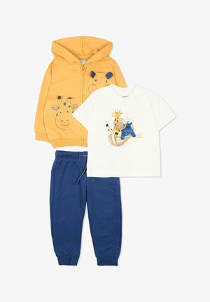 Żółta bluza z kapturem i zamkiem błyskawicznym z nadrukiem twarzy żyrafy, biała koszulka z nadrukiem zwierząt na łódce oraz niebieskie spodnie dresowe ze ściągaczem.