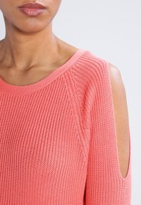 Sweter dzianinowy z odkrytymi ramionami w kolorze koralowym, z teksturowanym wzorem, ściągaczem przy dekolcie i szerokimi rękawami, zapewniający luźny krój i lekkie uczucie.