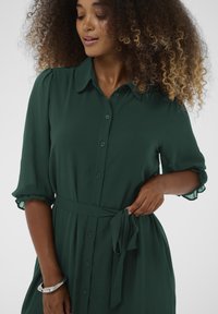 Tume roheline nööbiga kleit lühikeste volangkäistega ja vöödiga taljes. Valmistatud kergest kangast, millel on sile tekstuur.