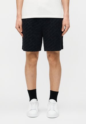 Personne portant un short noir texturé, des chaussettes noires, des baskets blanches et une chemise blanche, debout devant un fond clair uni.