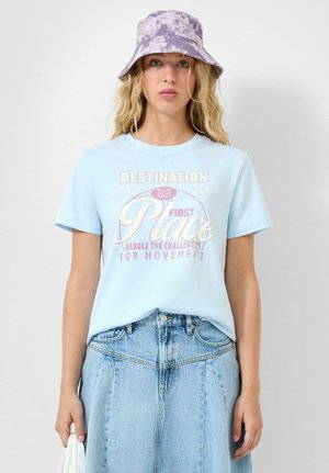 Jonge vrouw met lang blond haar draagt een paarse tie-dye bucket hat, lichtblauw T-shirt met opdruk en een hoogwater denim rok, en houdt een tas vast.