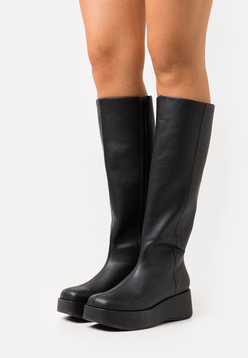 Botas negras, hasta la rodilla, hechas de cuero liso con una suela de plataforma gruesa y cremallera lateral. Diseño simple y moderno sin patrones visibles.