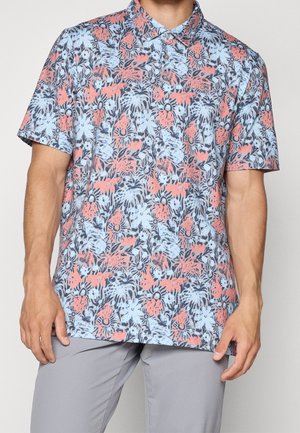 Homme portant une chemise à manches courtes boutonnée avec un motif floral tropical corail et bleu clair, associée à un pantalon gris clair.