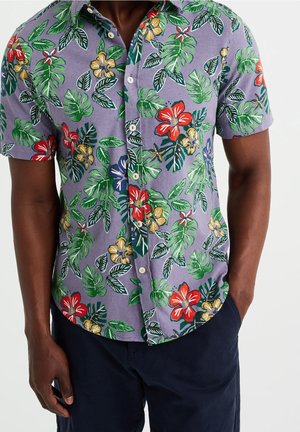 Chemise à manches courtes avec un fond violet, des motifs floraux vibrants en rouge, jaune et vert, avec une coupe décontractée et un tissu léger.