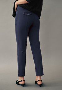 Pantaloni navy, croiți pe corp, cu un fit slim, având un buzunar pe spate cu nasture, cusături laterale și tăietură la glezne, purtați împreună cu pantofi cu toc negri.
