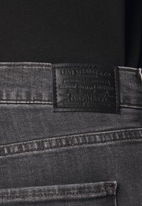 Grå denimjeans med en svart läderlapp med upphöjd text. Inkluderar sömnadsdetaljer och en vanlig fickdesign.