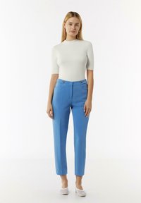 Pantalons taillés bleus avec une texture lisse, dotés de poches avant et de détails en métal doré. Assortis à un haut blanc ajusté à manches courtes.