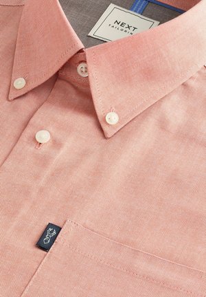 Chemise corail clair avec un tissage texturé, col boutonné et boutons couleur crème. Présente une étiquette bleu marine sur la poche.