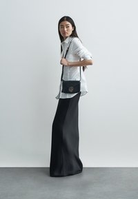 Fekete keresztpántos táska arany logóval, fémes díszítéssel és sima tapintású felülettel. Fehér, gombos blúzzal és testhezálló szoknyával párosítva.