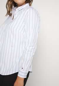 Hvid skjorte med lyseblå striber, krave, knaplukning foran, lange ærmer, brystlomme med "T.H." broderi og logoaccent på ærmet.