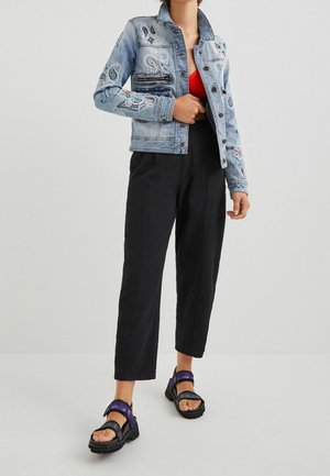 Personne portant une veste en jean brodée bleu clair, un haut rouge, un pantalon court noir et des sandales noires avec des lanières violettes.