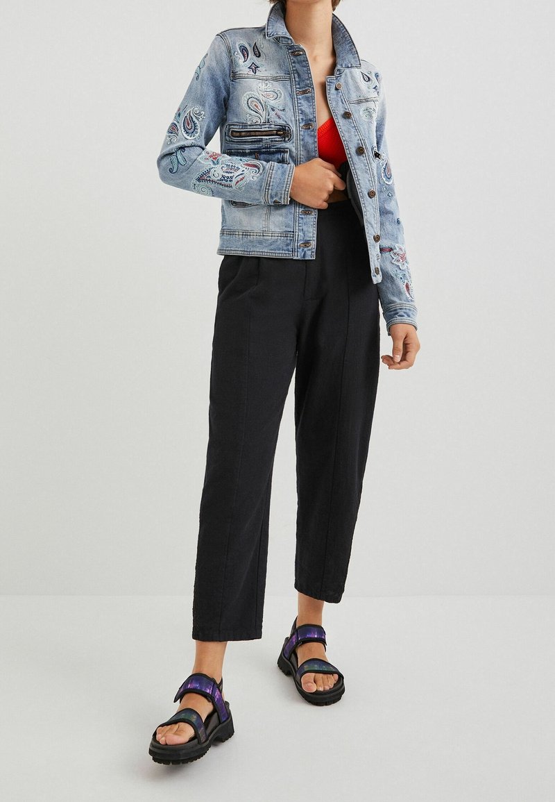 Personne portant une veste en jean brodée bleu clair, un haut rouge, un pantalon court noir et des sandales noires avec des lanières violettes.