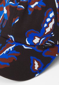 Pet met een gebogen rand, gemaakt van stof met een zwarte achtergrond en blauwe en bruine bloemmotieven. Inclusief een klein label aan de zijkant.