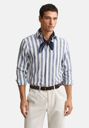 Homme portant une chemise à rayures verticales bleu et blanc, un foulard bleu marine autour du cou, un pantalon beige clair et une ceinture sombre à motifs, debout, les mains détendues.