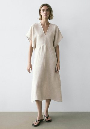 Donna in piedi contro un muro bianco che indossa un abito midi beige con scollo a V, maniche larghe e sandali neri infradito.