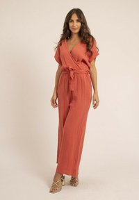 Combinaison de couleur rouille avec un tissu texturé, design enveloppant, manches courtes et une ceinture assortie. Associée à des escarpins dorés à bride.