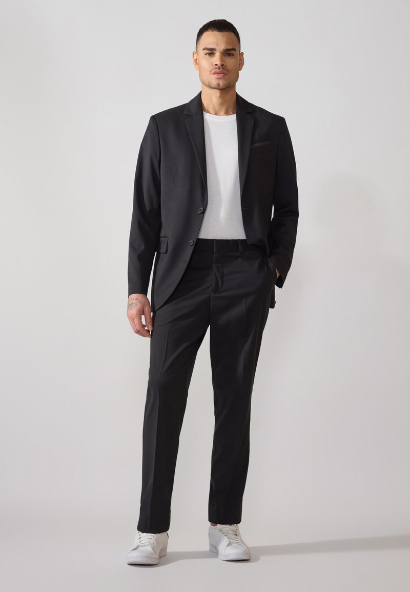 Costum negru cu un design slim-fit, sacou cu un singur nasture și pantaloni drepți; asortat cu un tricou alb și adidași albi.