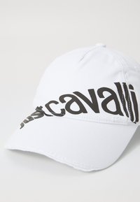 Balts beisbola cepure ar izliekto purngalu, kurai pa priekšu ir izcila melna "justcavalli" logo diagonāli.