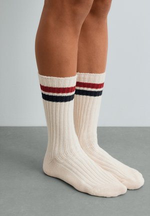 Jambes portant des chaussettes côtelées crème avec des rayures rouges et noires près du haut, debout sur un sol gris contre un mur pâle.