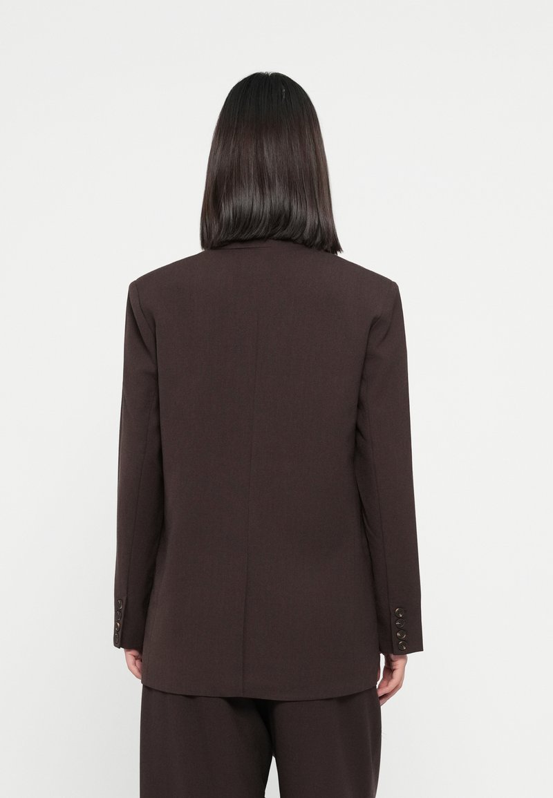 Brązowa, dopasowana marynarka o usztywnionym kroju, z klapami i guzikami przy mankietach. Gładki materiał, minimalne detale, rozporek z tyłu dla lepszego dopasowania.