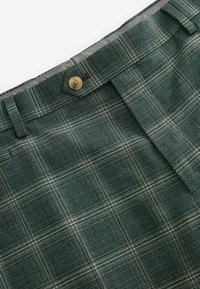 Karierte Hose aus dunkelgrünem Stoff mit beigen und weißen Überprüfungslinien, einem Knopfverschluss und einem glatten Innenfutter.