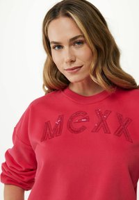 Sudadera roja con el texto "MCXX" adornado con lentejuelas. Presenta un escote redondo y mangas enrolladas, confeccionada con una tela suave y texturizada.