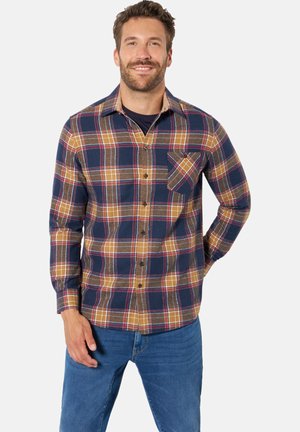 Man smiling wearing a blue, brown, and yellow plaid button-up shirt with blue jeans, one hand in pocket and white background.