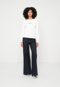 Witte sweatshirt met blauwe tekstlogo, gecombineerd met donkere, wijde spijkerbroek. De voeten van het model zijn in puntige zilveren hakken. Eenvoudige achtergrond.