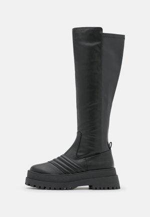Botte haute en cuir noir avec semelle épaisse à crampons, détails avant matelassés et languette au talon, montrée de profil sur fond blanc.