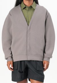 Šedá oversized mikina s V-výstřihem, žebrovanými manžetami a spodním lemem, vrstvená přes zelenou košili s knoflíky, kombinovaná s černými šortkami.