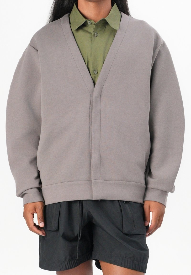 Šedá oversized mikina s V-výstřihem, žebrovanými manžetami a spodním lemem, vrstvená přes zelenou košili s knoflíky, kombinovaná s černými šortkami.