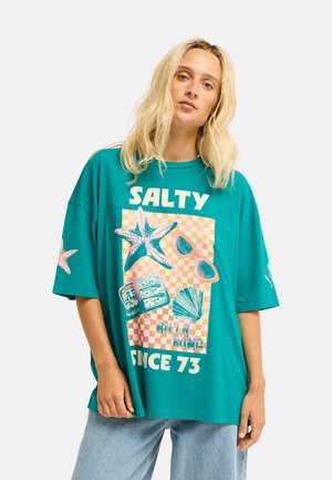 Blonde vrouw draagt een oversized teal T-shirt met een zeester, zonnebril, schelp, blik vis grafiek en tekst "Salty Since 73" en lichtblauwe spijkerbroek.