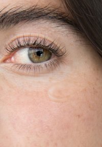 Nærbilde av et øye med brun iris og lange øyevipper, omgitt av lys hud med fregner. En liten dråpe klar væske hviler på kinnet.