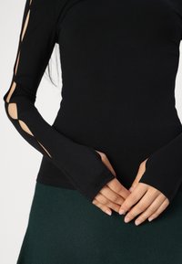 Top negru cu mâneci lungi, având detalii decupate pe brațe, realizat dintr-un material moale. Mânele dispun de găuri pentru degete, completând o fustă verde.