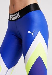 Närbild av blå, vita och neon gula Puma leggings med svart midjeband som har Puma-logotypen på höften och midjan.